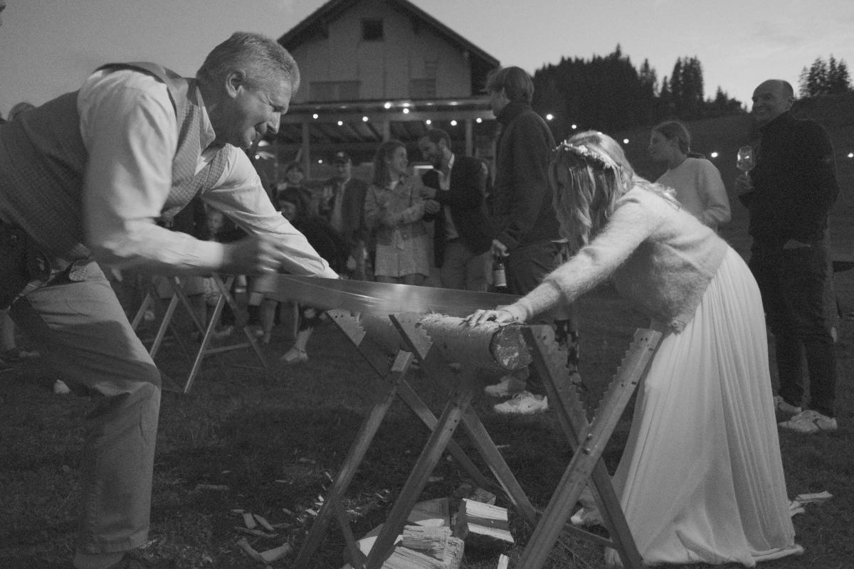 Braut und Gast sägen gemeinsam einen Baumstamm, umringt von lachenden Hochzeitsgästen vor einem Stall mit Lichterkette: Hochzeit mit MicheleKocht in der Schweiz