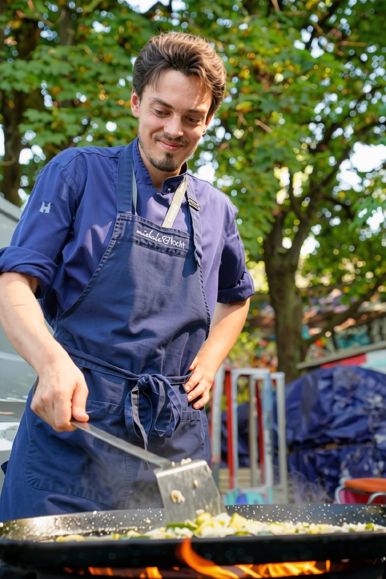 Chef in a blue apron stirring the large pan over an open flame: live cooking with MicheleKocht, Zürich