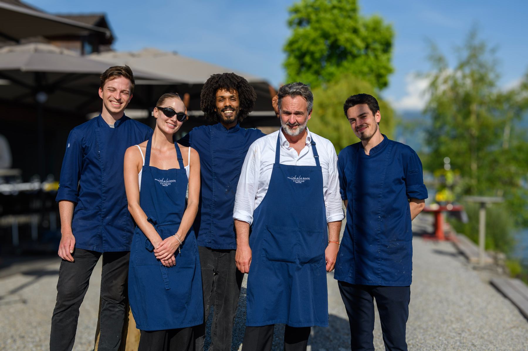 Michele mit seinem Team in blauen Kochjacken und Schürzen am See: MicheleKocht Catering Zürich